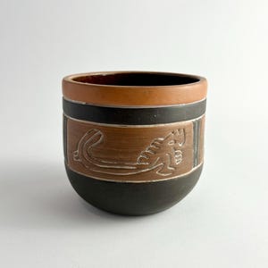 May include: A handcrafted ceramic cup with a unique design. The cup features a brown exterior with a black band and a detailed relief of a creature. The rim is a lighter brown, and the interior is dark brown.