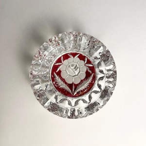 May include: A circular clear glass ashtray featuring a faceted, cut-glass rim and a central red inlay. The red center displays a white etched floral design with a central flower and two leaves, creating a distinct contrast against the clear glass.