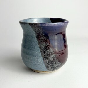 May include: A handmade ceramic tumbler with a unique color scheme. The cup features a blend of blue, purple, and maroon hues. The design includes a diagonal color split, adding visual interest to the handcrafted pottery.