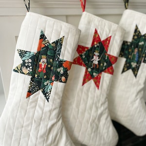 Puede incluir: Tres calcetines de Navidad blancos acolchados con diseños de patchwork en forma de estrella. Los calcetines están colgados de ganchos y presentan diferentes patrones, incluyendo un diseño verde y dorado con un cascanueces, un diseño rojo y dorado con un cascanueces y un diseño negro y dorado con una figura.