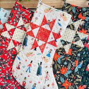 May include: Three red and white quilted Christmas stockings with different patterns. The stocking on the left has a red and white star pattern, the middle stocking has a white background with a red star pattern and a variety of Christmas characters, and the stocking on the right has a black background with a green, red, and gold floral pattern.