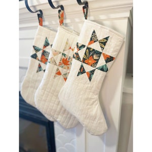 Peut inclure: Trois chaussettes de Noël blanches matelassées avec un motif en étoile éclatante en tissu orange, vert et noir. Les chaussettes sont accrochées à une cheminée blanche.