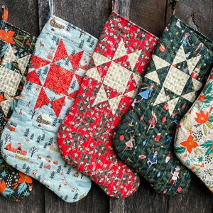 May include: Six Christmas stockings made of fabric with a patchwork design. The stockings are decorated with a variety of festive themes, including stars, snowflakes, and poinsettias. The stockings are hung on a wooden wall.