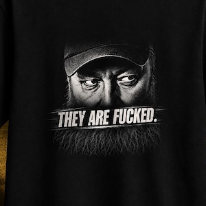 May include: A black short-sleeved t-shirt featuring a graphic print of a bearded person wearing a baseball cap. The text "THEY ARE FUCKED." is printed in white capital letters across the centre of the chest, positioned over the beard area.