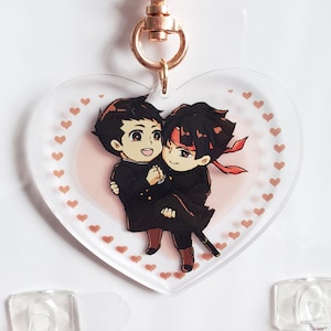 May include: A heart-shaped acrylic keychain with a pink background and a white border. The keychain features two anime characters, one with black hair and a red headband, holding the other character with brown hair. The background is filled with small white hearts.