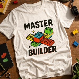 May include: White t-shirt with the words "MASTER BUILDER" in black, featuring a colorful graphic of stacked toy blocks. The blocks are red, yellow, green, and blue. The shirt is on a wooden surface.