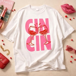Puede incluir: Camiseta blanca con la inscripción rosa "CIN CIN" y dos copas de vino tinto brindando. La camiseta es de estilo casual.