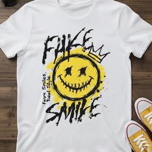 Puede incluir: Camiseta blanca con un diseño gráfico que presenta una carita sonriente amarilla con una corona, rodeada por las palabras "FAKE SMILE" y "Faux Smiles, Real Style" en negro. La camiseta se combina con zapatillas amarillas.
