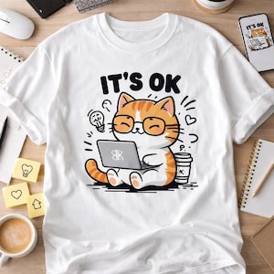 Puede incluir: Camiseta blanca con un diseño de gato de dibujos animados. El gato, con pelaje naranja y blanco, lleva gafas y usa un portátil. El texto "IT'S OK" está encima del gato. Una taza de café y una bombilla también están en el diseño.