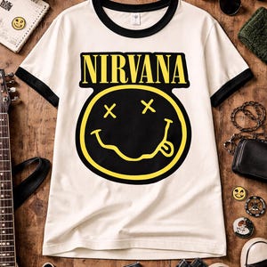 May include: Cream-colored ringer t-shirt with black trim featuring the word "NIRVANA" in yellow block letters above a yellow smiley face with black eyes and tongue. The image includes a guitar, headphones, and other accessories.