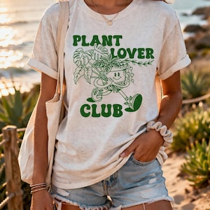 May include: A light beige t-shirt with the text "PLANT LOVER CLUB" in green. The graphic features a smiling plant pot character holding a bouquet of plants. The shirt is paired with denim shorts.