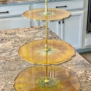 May include: A three-tiered serving stand with gold metal accents. Each tier is a round, scalloped, golden-brown resin tray with a glittery finish.