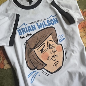 May include: White t-shirt with black trim on the collar and sleeves. The shirt features a cartoon illustration of a person with the text "PRESIDENT OF THE BRIAN WILSON fan club" in blue.