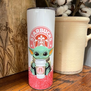 Puede incluir: Un vaso Starbucks blanco con el logotipo rosa de Starbucks y una imagen de dibujos animados de Grogu sosteniendo una taza de café Starbucks. La parte inferior del vaso tiene un diseño de purpurina roja.