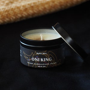 Peut inclure: Une boîte de bougie noire avec une étiquette blanche qui indique "Mythic Mori Oni King coconut sandalwood musk - strength net wt. 3oz". La bougie est allumée et le couvercle est ouvert.