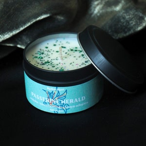 May include: A teal blue candle with a black lid and a white label that says "Mythic Mort Passerin Herald" with a bird illustration. The candle is filled with white wax and green glitter.