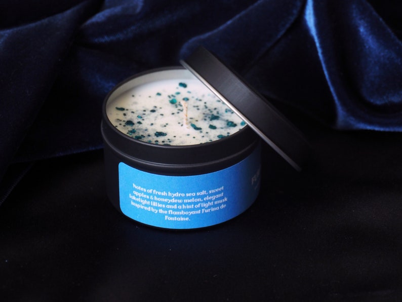 May include: A black candle tin with a blue label that reads "Notes of fresh hydro sea salt, sweet apples & honeydew melon, elegant night lilies and a hint of light musk inspired by the flamboyant Furia de Fontaine."