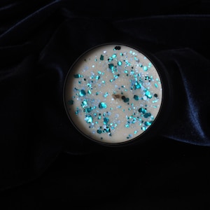 May include: A round candle with a beige wax base and a layer of teal and silver glitter on top. The candle is in a black container and is sitting on a dark blue velvet surface.