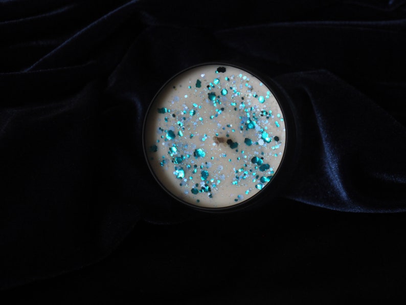 May include: A round candle with a beige wax base and a layer of teal and silver glitter on top. The candle is in a black container and is sitting on a dark blue velvet surface.