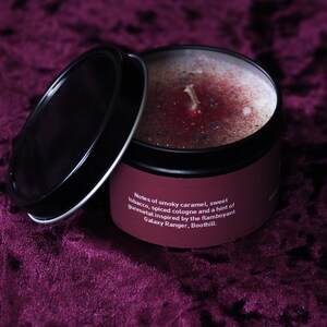 May include: A black tin candle with a red and silver wax top. The label reads "Notes of smoky caramel, sweet tobacco, spiced cologne and a hint of gunmetal inspired by the flamboyant Galaxy Ranger, Boothill."