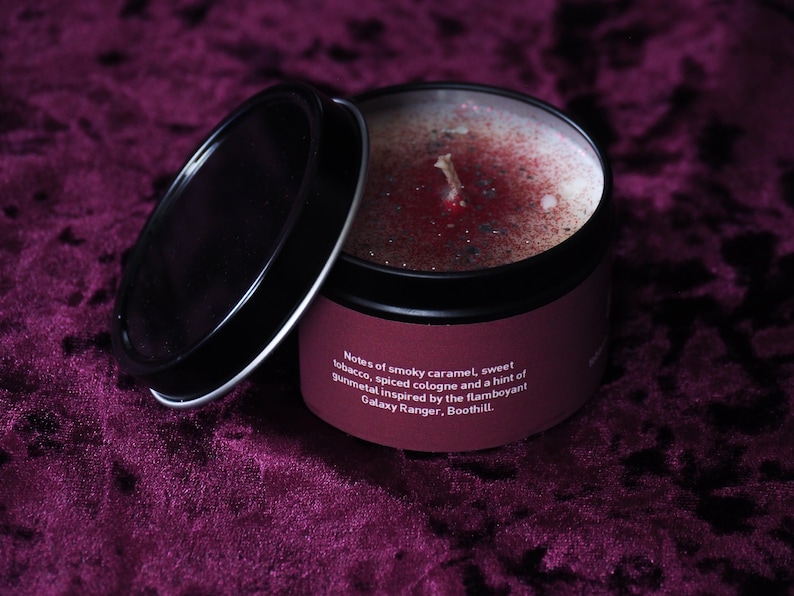 May include: A black tin candle with a red and silver wax top. The label reads "Notes of smoky caramel, sweet tobacco, spiced cologne and a hint of gunmetal inspired by the flamboyant Galaxy Ranger, Boothill."