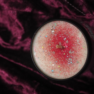 May include: A white candle with red glitter and a black wick in a black glass jar.