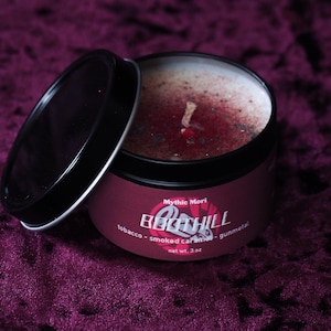 May include: A black candle tin with a red and white label that reads "Mythic Mori Beech Hill tobacco - smoked caramel - gunmetal net wt. 3 oz". The candle is lit and the wax is a dark red colour.