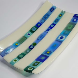 Puede incluir: Un plato rectangular de vidrio blanco con tres patrones de rayas azules, verdes y amarillas. El plato es ligeramente curvo y tiene un acabado brillante.