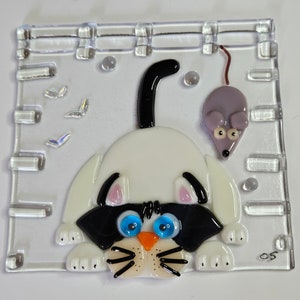Puede incluir: Un gato blanco con orejas negras y una cola negra está mirando a un ratón morado con una cola roja. El gato tiene ojos azules y una nariz naranja. El gato y el ratón están hechos de vidrio y están sobre un fondo de vidrio transparente. También hay pequeñas mariposas iridiscentes en el fondo.