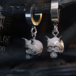 Può includere: Orecchini a cerchio argentati con ciondoli a forma di teschio. Ogni teschio è bianco con sfumature grigie e piccole corna. Gli orecchini sono appesi a un ramo scuro. Il testo "FRIGHT LIGHT STUDIO" è visibile sullo sfondo.