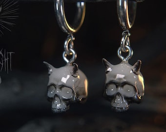 May include: A pair of silver hoop earrings with skull charms. Each skull is painted in a light gray color with dark gray accents and small horns. The earrings are suspended from a dark branch. The text "FRIGHT LIGHT STUDIO" is visible.