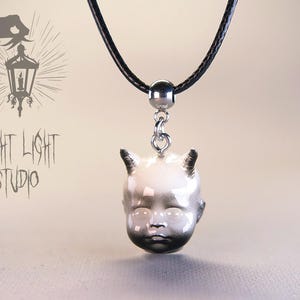 Peut inclure: Un collier pendentif avec une tête de poupée bébé détaillée, ornée de cornes et d'un dégradé de gris. La tête est suspendue à un cordon noir avec un fermoir en métal argenté. Le texte "FRIGHT LIGHT STUDIO" est visible en arrière-plan.