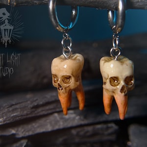 Könnte beinhalten: Ein Paar silberne Creolen mit Anhängern in Form von Zähnen mit Totenkopfgesichtern. Die Zähne sind cremefarben mit orangefarbenen Wurzeln. Die Ohrringe werden vor einem dunklen, strukturierten Hintergrund präsentiert. Der Text "FRIGHT LIGHT STUDIO" ist sichtbar.