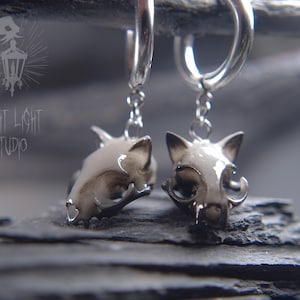 May include: A pair of silver hoop earrings with a small, detailed, white cat skull charm hanging from each hoop.
