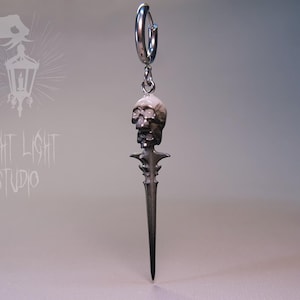 Pode incluir: Brinco prateado com um pendente de caveira e punhal. A caveira é detalhada e o punhal tem um cabo ornamentado. O brinco pende de um aro. O texto "FRIGHT LIGHT STUDIO" é visível.