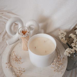 May include: A white candle with three wicks sits on a white tray with dried white flowers and a glass jar of matches. The tray is on a white fabric surface.