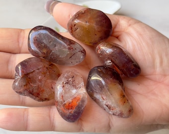 May include: Six tumbled stones of clear quartz with red and brown inclusions. The stones are smooth and polished, and they have a variety of shapes and sizes.