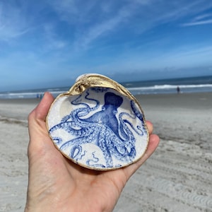 May include: A decorative seashell with a blue octopus design inside. The shell's rim is painted gold. The background shows a beach with blue sky and ocean.