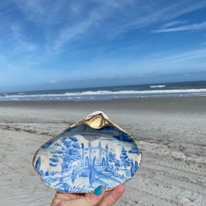 May include: A hand-painted seashell trinket dish with a blue and white landscape design. The interior features a detailed scene of a building and trees, with a gold-painted rim. The dish is held against a backdrop of a beach and blue sky.