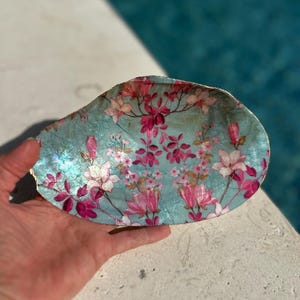 以下が含まれることがあります： 手描きの装飾的な貝殻の皿。 ターコイズブルーの背景にピンクと白の花柄が描かれています。縁は金でトリミングされています。 ジュエリーや小物を入れるのに最適です。