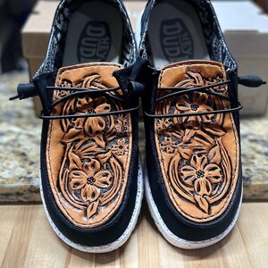 May include: A pair of black canvas shoes with a brown leather floral design. The shoes have a white sole and the text "Hey Dud" is printed on the inside of the shoes.