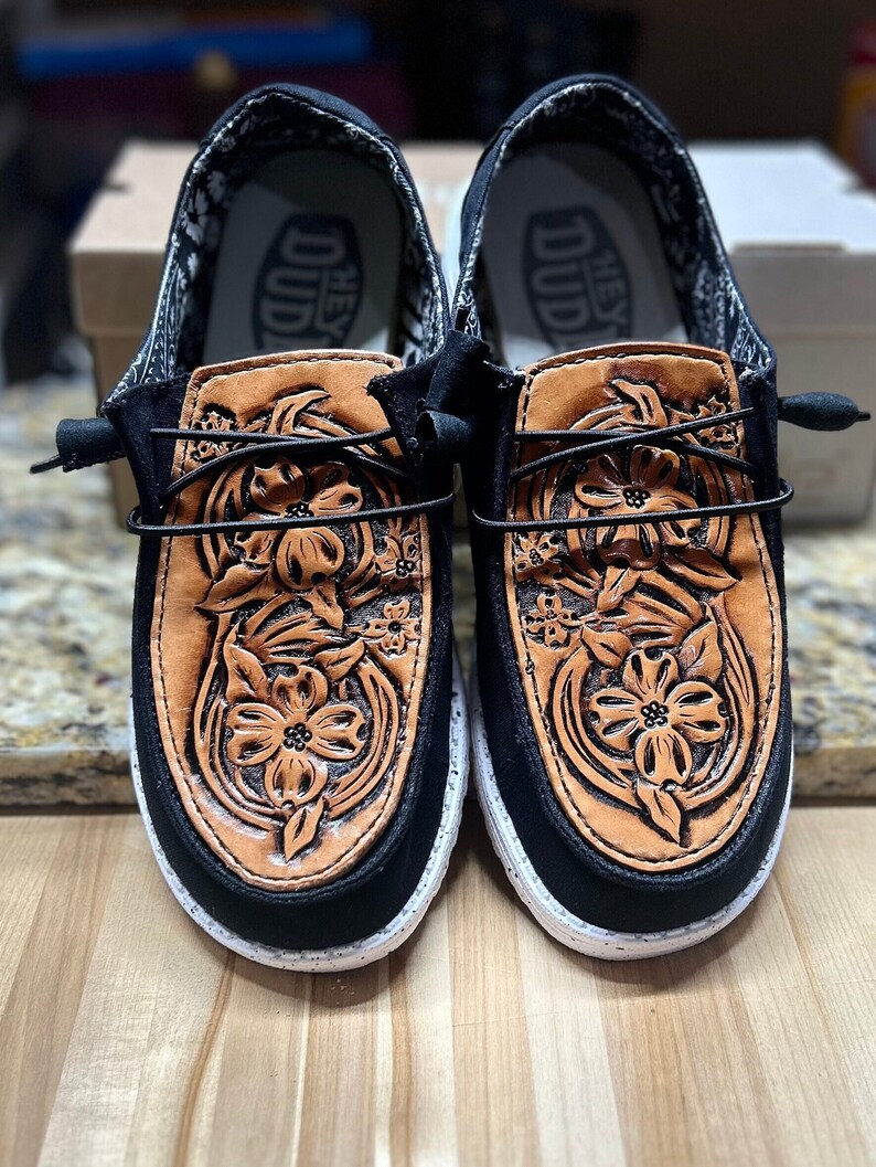 May include: A pair of black canvas shoes with a brown leather floral design. The shoes have a white sole and the text "Hey Dud" is printed on the inside of the shoes.