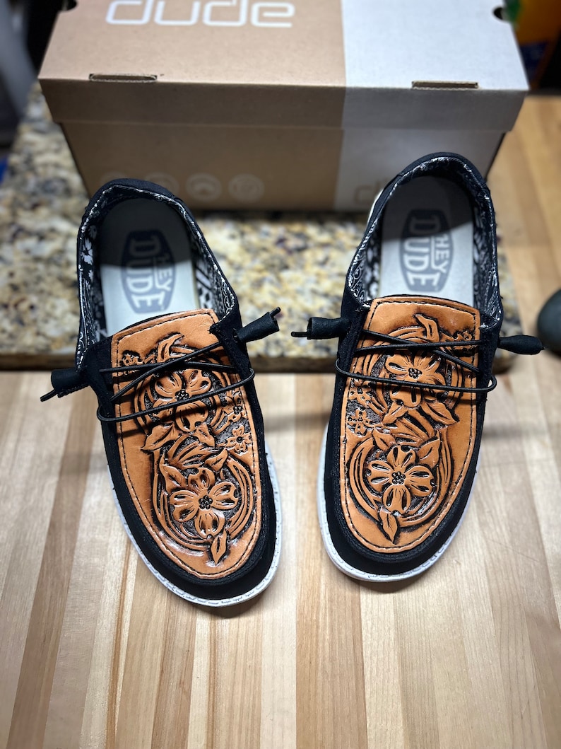 May include: A pair of black canvas shoes with brown leather floral embroidery. The shoes have a white sole and the text "Hey Dude" is visible on the inside of the shoes.