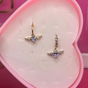 May include: A pair of gold-toned earrings, each featuring a heart-shaped purple gemstone with delicate wing accents. One earring is a simple hoop, while the other has a row of small, clear stones above the heart. The earrings are displayed in a pink heart-shaped box.