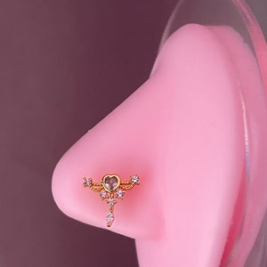 May include: Gold nose ring with a heart-shaped centre stone and smaller stones on the sides. The ring is on a pink display stand.