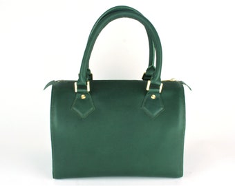 Puede incluir: Un bolso de mano verde oscuro con dos asas curvas y un cuerpo rectangular. El bolso presenta herrajes dorados en las asas y un cierre de cremallera. El bolso está hecho de un material liso y tiene una forma estructurada.