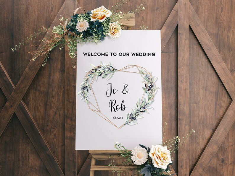 May include: A white wedding sign with a gold geometric heart frame and olive branch wreath. The sign reads "Welcome to our wedding" and "Jo & Rob" with the date "02.04.22".