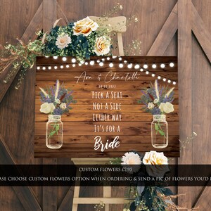 May include: A rustic wooden sign with a white background and the text "Pick a seat, not a side, either way it's for a bride". The sign is decorated with mason jars filled with flowers and a string of lights.