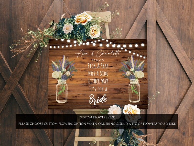 May include: A rustic wooden sign with a white background and the text "Pick a seat, not a side, either way it's for a bride". The sign is decorated with mason jars filled with flowers and a string of lights.