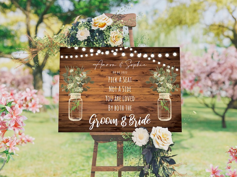 May include: A rustic wooden sign with a floral design and mason jars. The sign reads "Aaron & Sophie 09-03-2027 Pick a Seat Not a Side You Are Loved By Both The Groom & Bride".